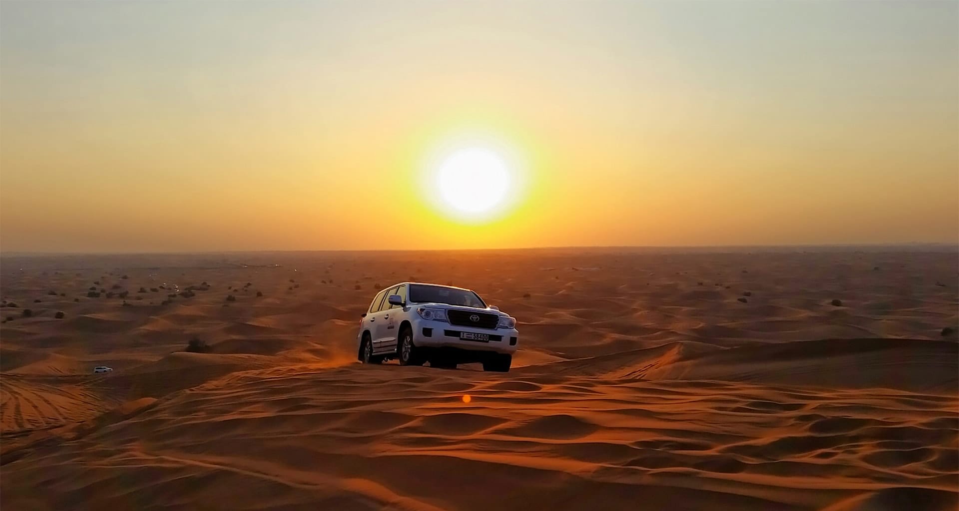 White SUV in Dubai desert at sunset, surrounded by sand dunes, capturing the thrill of dune bashing in a golden landscape.