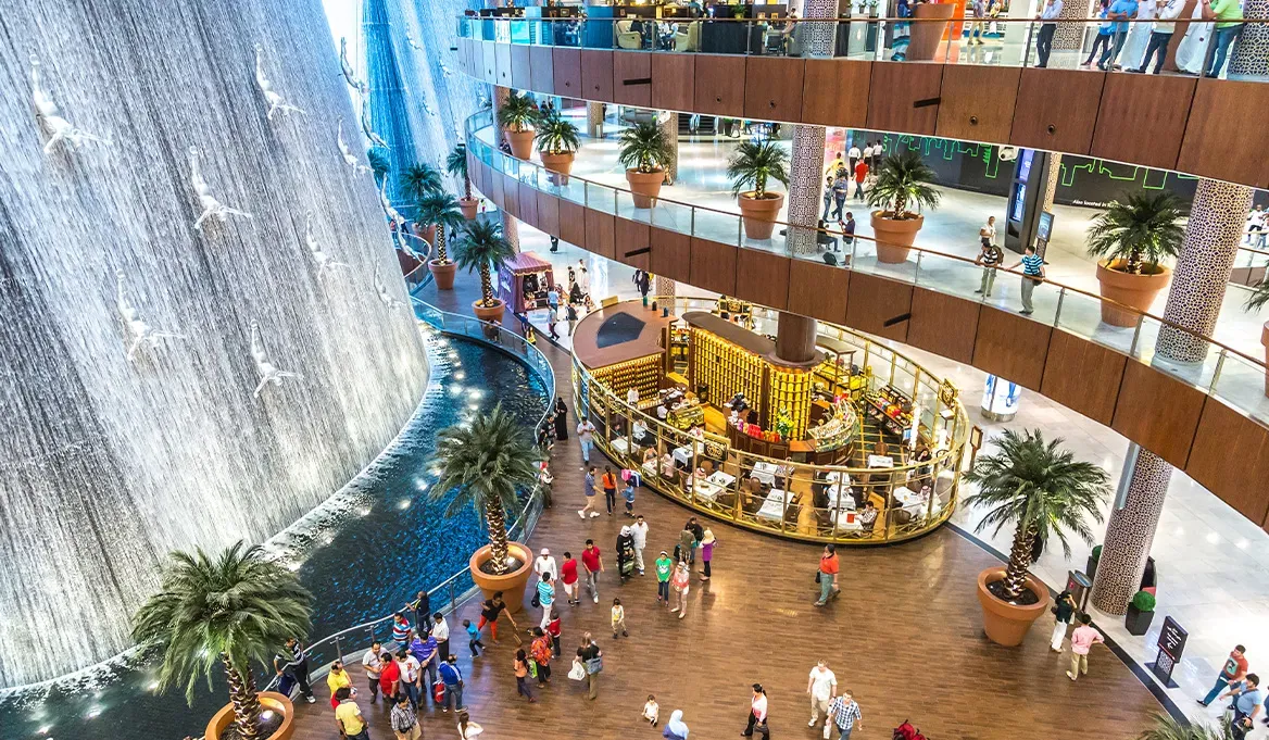 Dubai Mall interior with iconic waterfall, divers, palm trees, shops, and bustling crowds in a luxurious shopping setting.