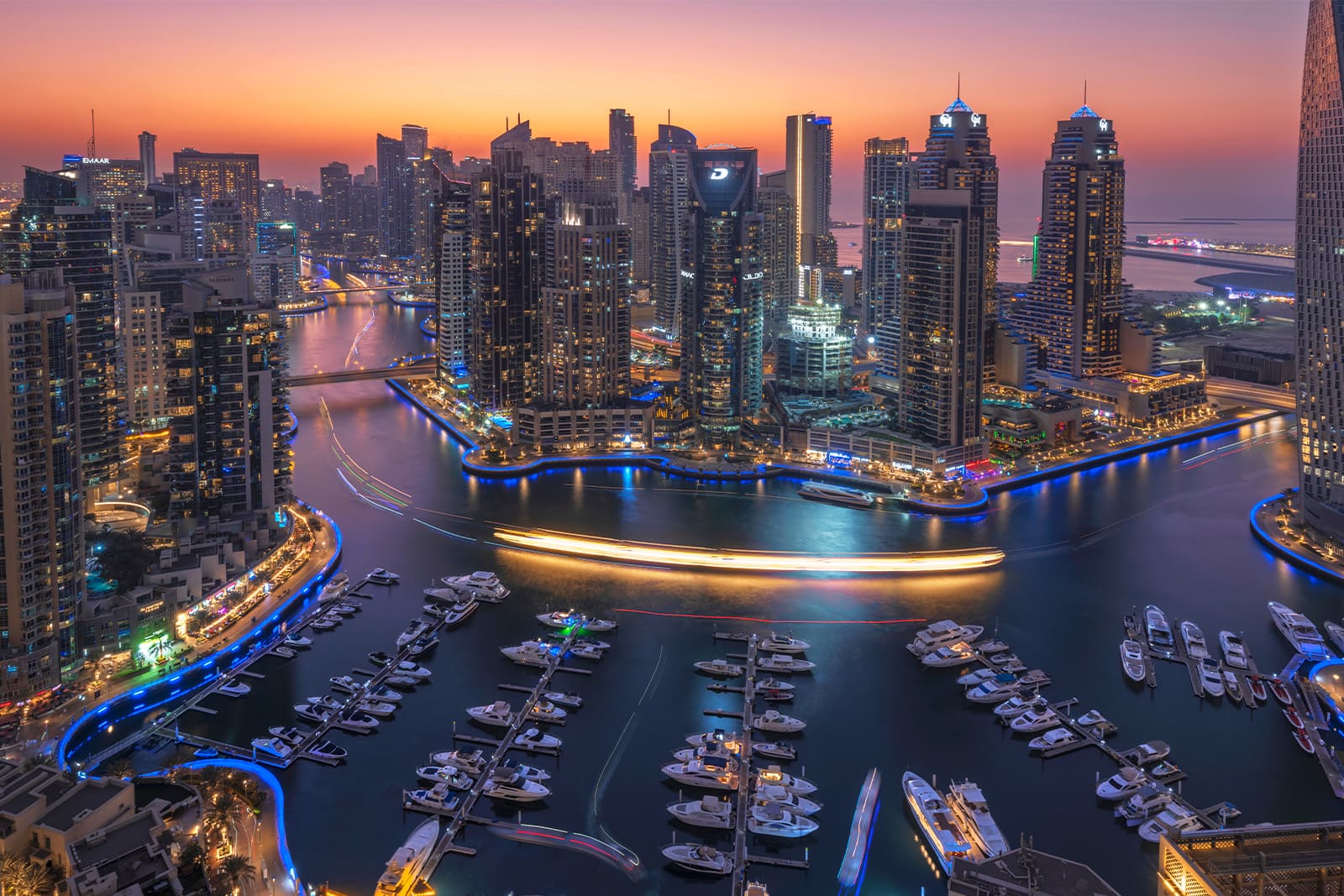 Dubai Marina skyline at sunset with illuminated skyscrapers, yachts in the marina, and vibrant city lights reflecting on the water.