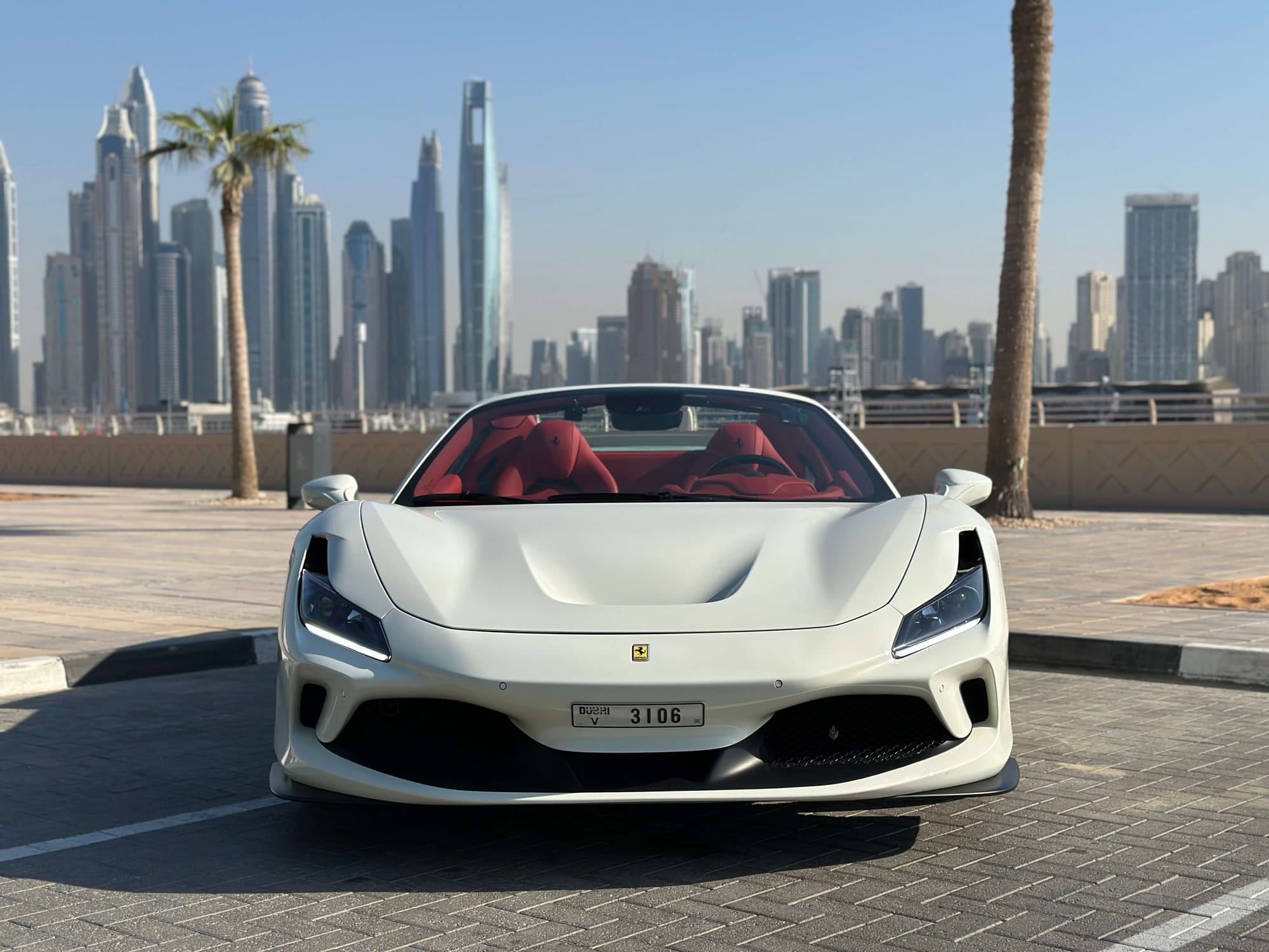 White Ferrari F8 Spider with red interior parked in Dubai with skyscrapers and palms.