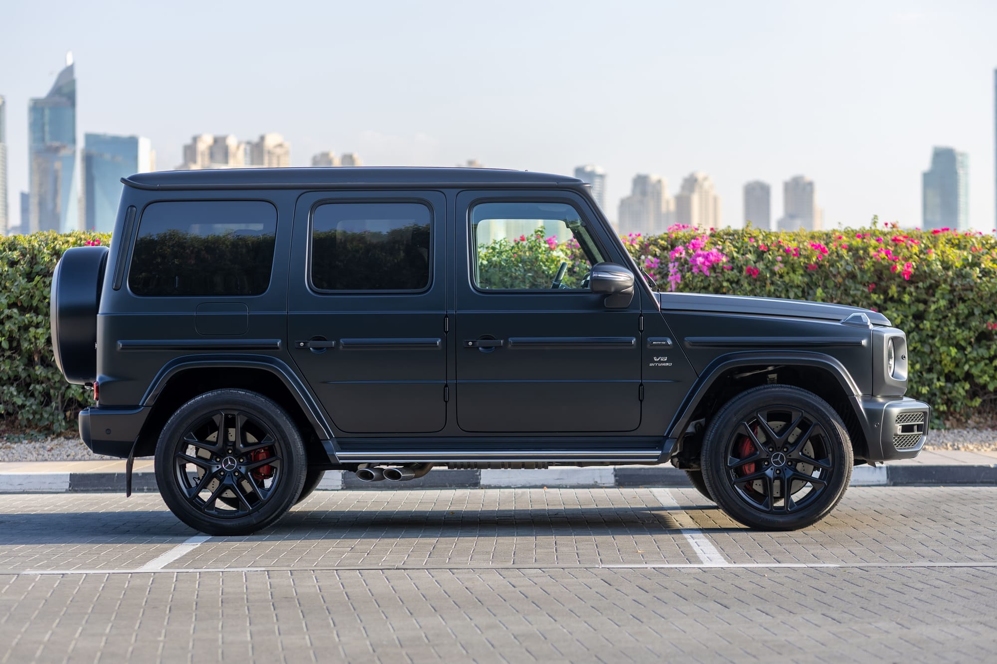 Black Mercedes-Benz G-Class AMG parked in Dubai with skyline and pink flowers.