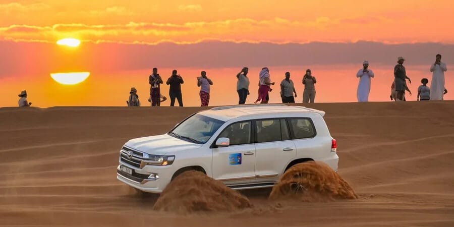 White SUV dune bashing in Dubai desert at sunset with tourists watching, highlighting tipping for safari guides.