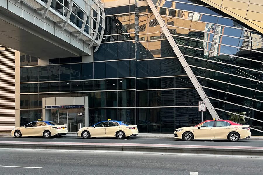 Cream-colored taxis parked outside Dubai Internet City Metro Station near glass building facade.