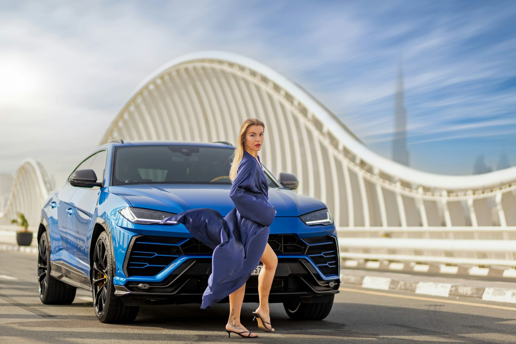 Blue Lamborghini Urus with woman posing on Dubai bridge near Burj Khalifa, symbolizing luxury car rental