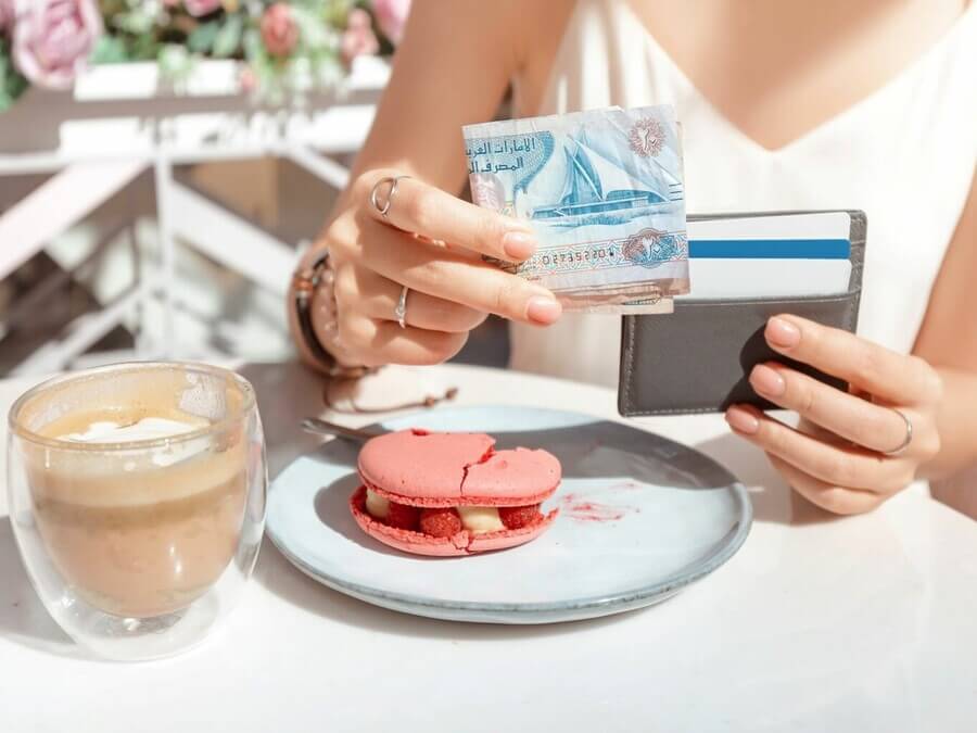 Tourist holding UAE dirham cash and card at cafe table with dessert and coffee, preparing to tip in Dubai.