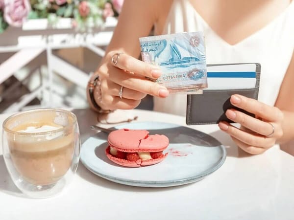 Tourist holding UAE dirham cash and card at cafe table with dessert and coffee, preparing to tip in Dubai.